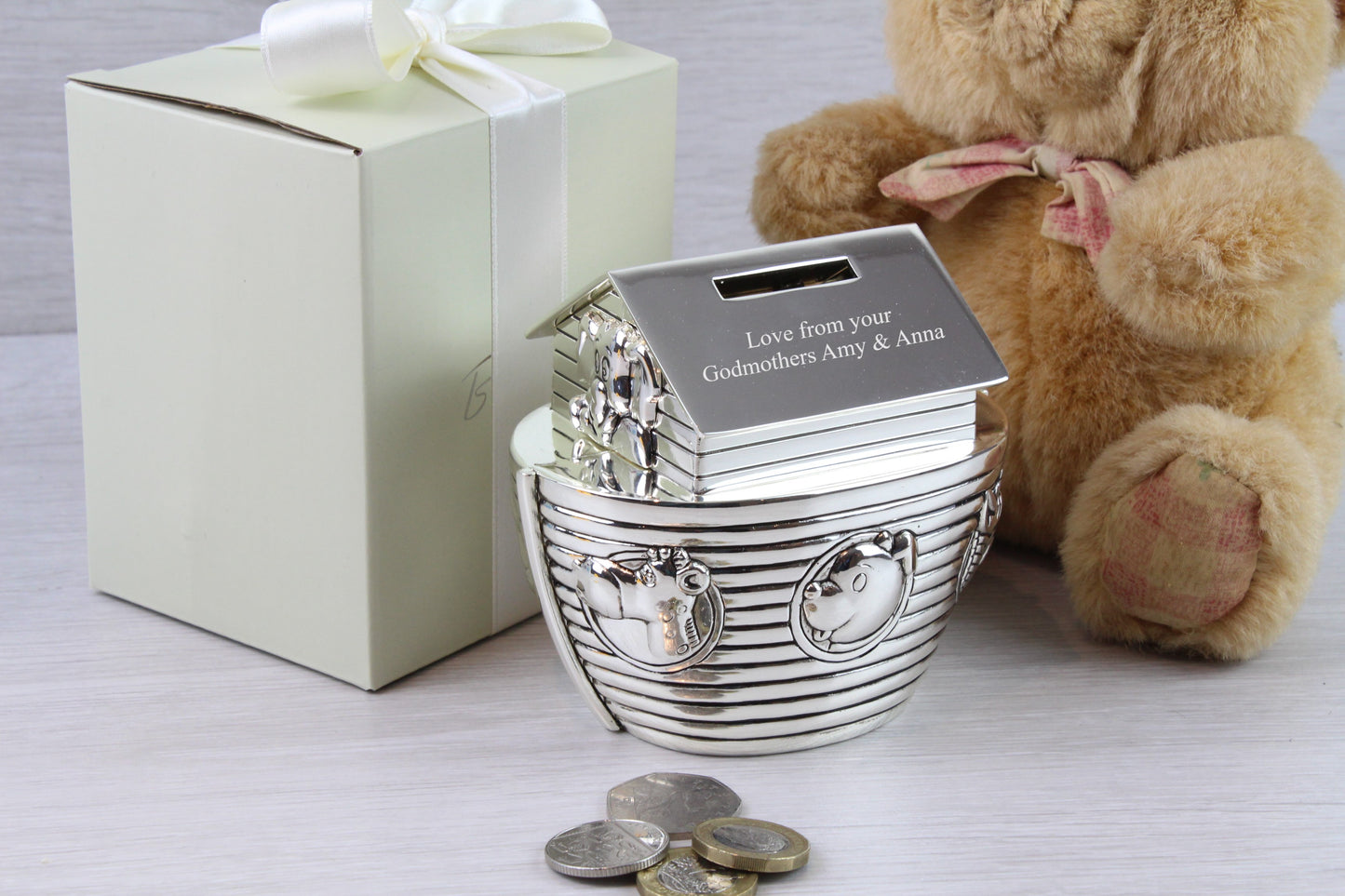 Personalised Noah's Ark Money Box that says "Love from your Godmothers Amy & Anna" with gift box, a brown teddy bear and some coins as props.