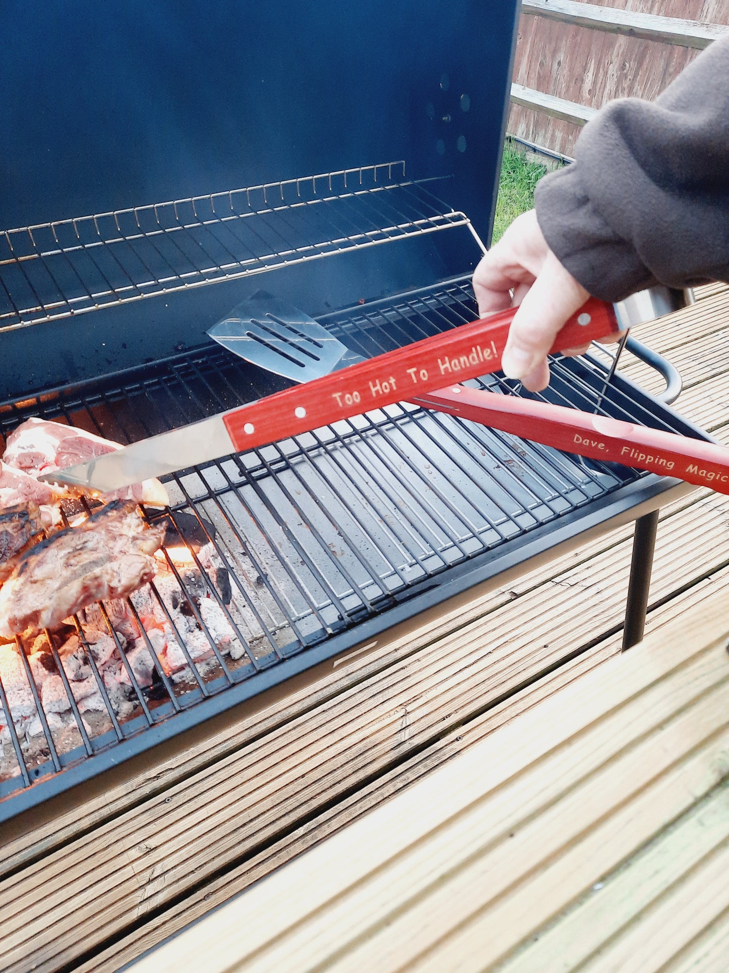 Grilling some pork using a personalised wooden handled tong that says "Too Hot To Handle!" and spatulad that says "Dave Flipping Magician"
