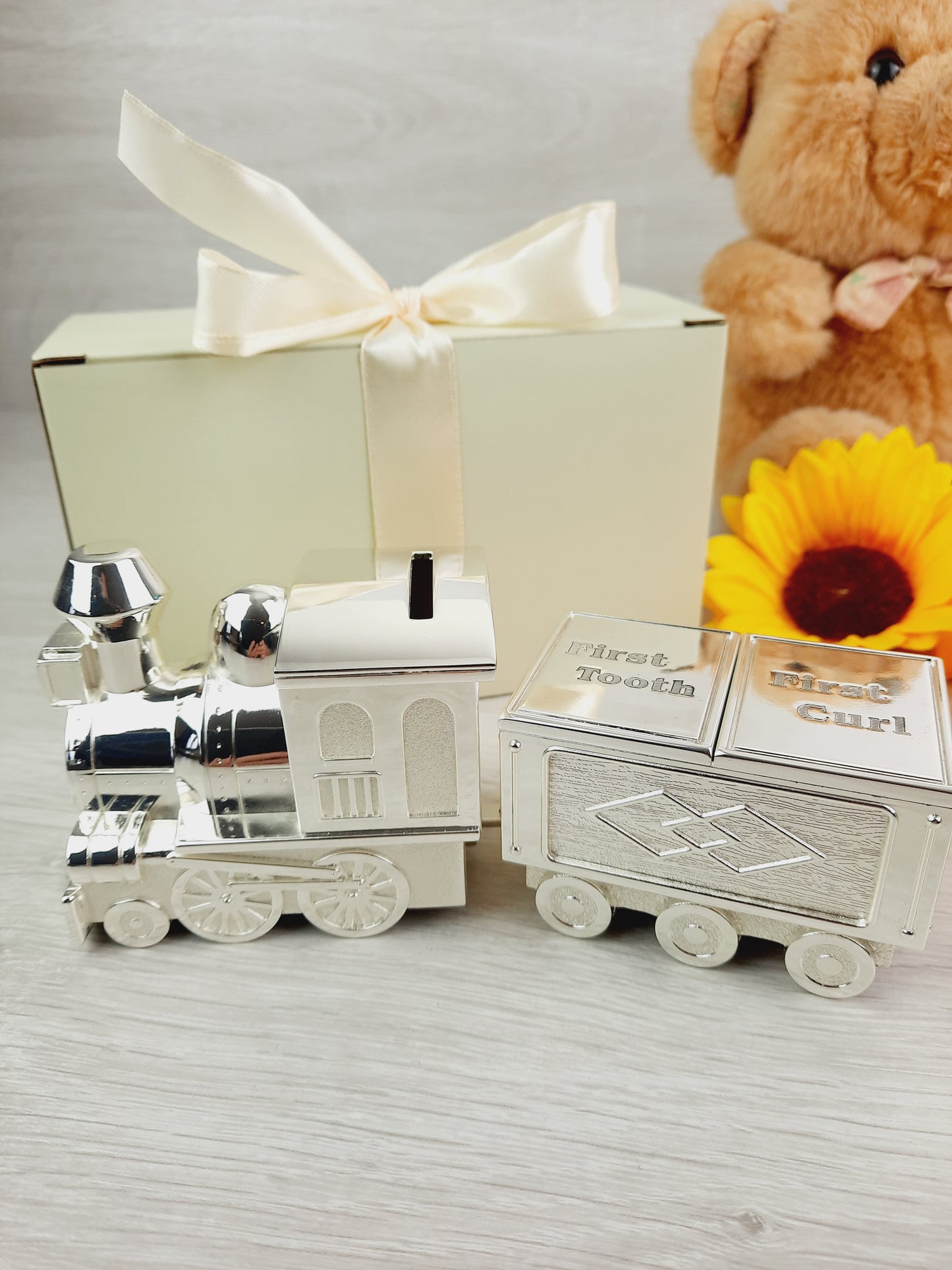 Personalised Engraved Money Box with Baby Tooth and Curl Train. There's a cream gift box with peach ribbon, a sunflower and brown teddy bear as props.