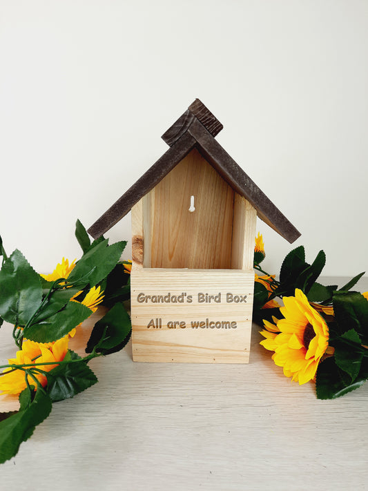 Personalised Bird Nesting Box says "Grandad's Bird Box All are welcome" with faux sunflowers surrounding it as props.