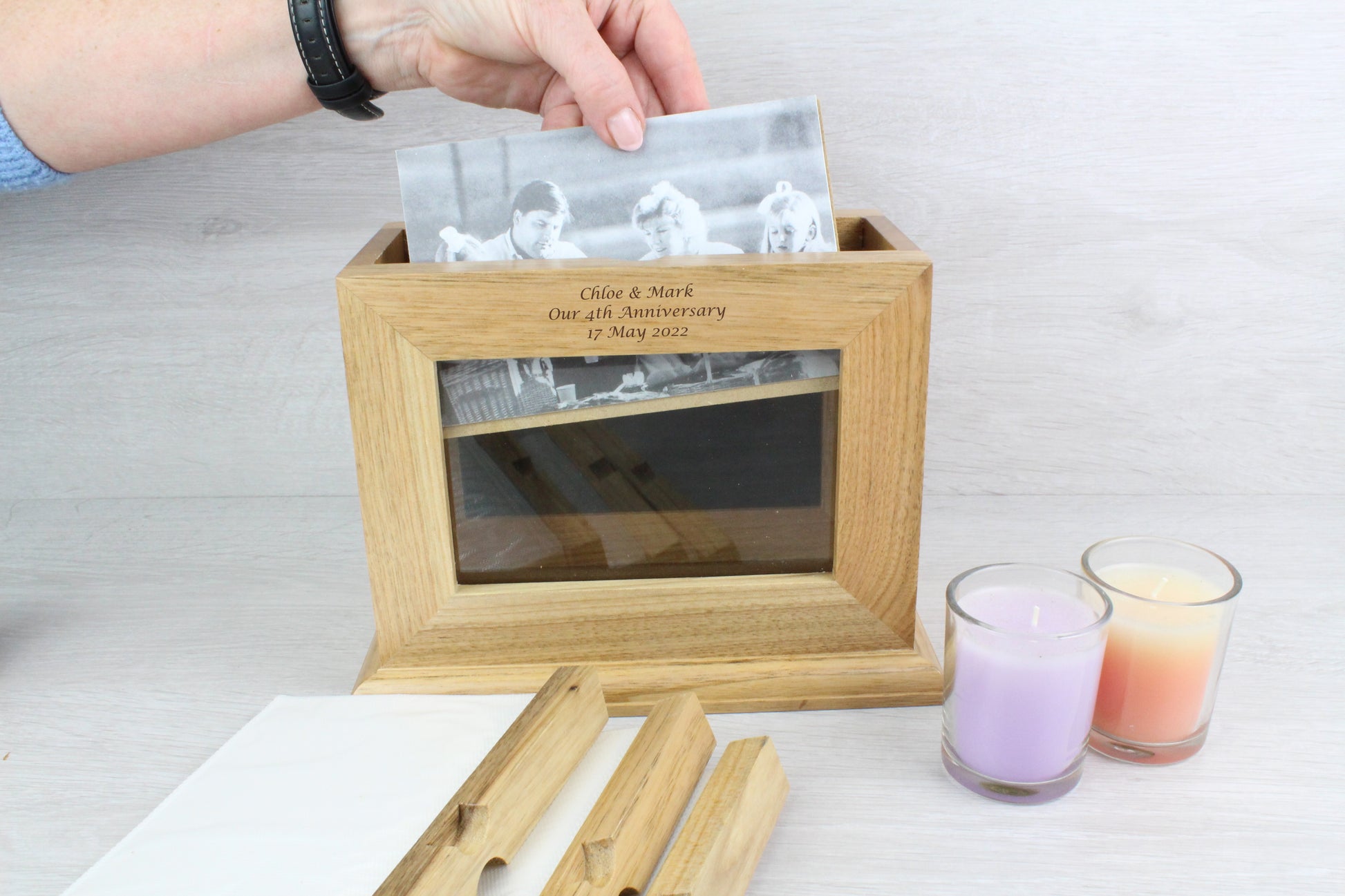 Demonstrating of how to insert the photo inside the ﻿Personalised Oak Wooden Photo Frame and Album Holder with a text engraving that says "Chloe & Mark Our 4th Wedding Anniversary 17 May 2022" Also, beside the wooden frame, there are two not lighted candles (colour purple and orange).