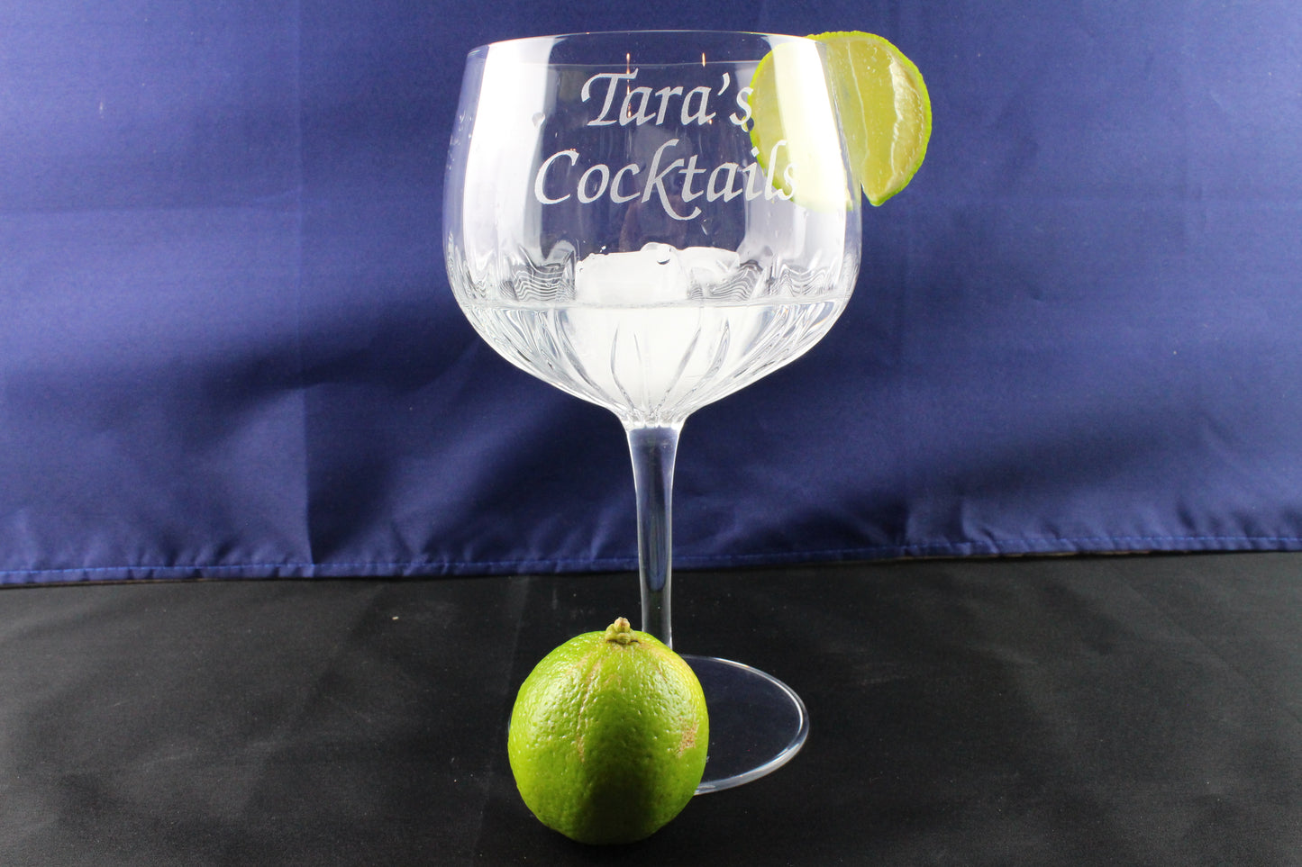 Personalised crystal cut gin glass with blue cloth background. A sliced lime is on the glass's mouth and 1 whole slime at the bottom of the glass.