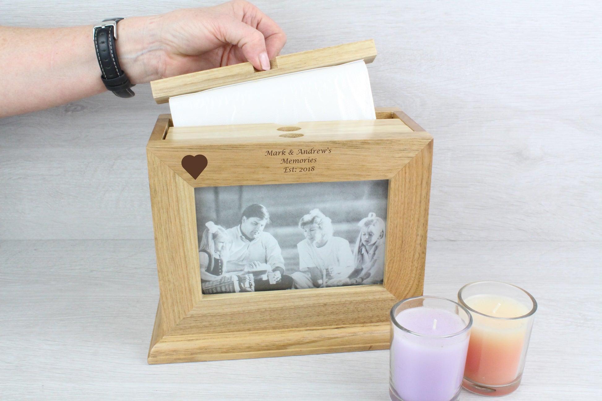 Demonstrating of how to insert the photo inside the ﻿Personalised Oak Wooden Photo Frame and Album Holder with names and a heart design that says "Mark & Andrews Memories Est. 2018". Also, beside the wooden frame, there are two not lighted candles (colour purple and orange).