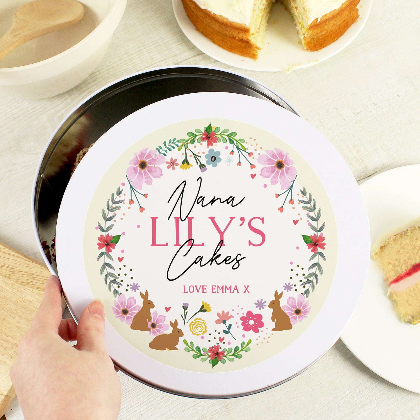 A hand is holding the tin cover of Personalised Floral Cake Tin that says "Nana LILY'D Cakes LOVE EMMA X"