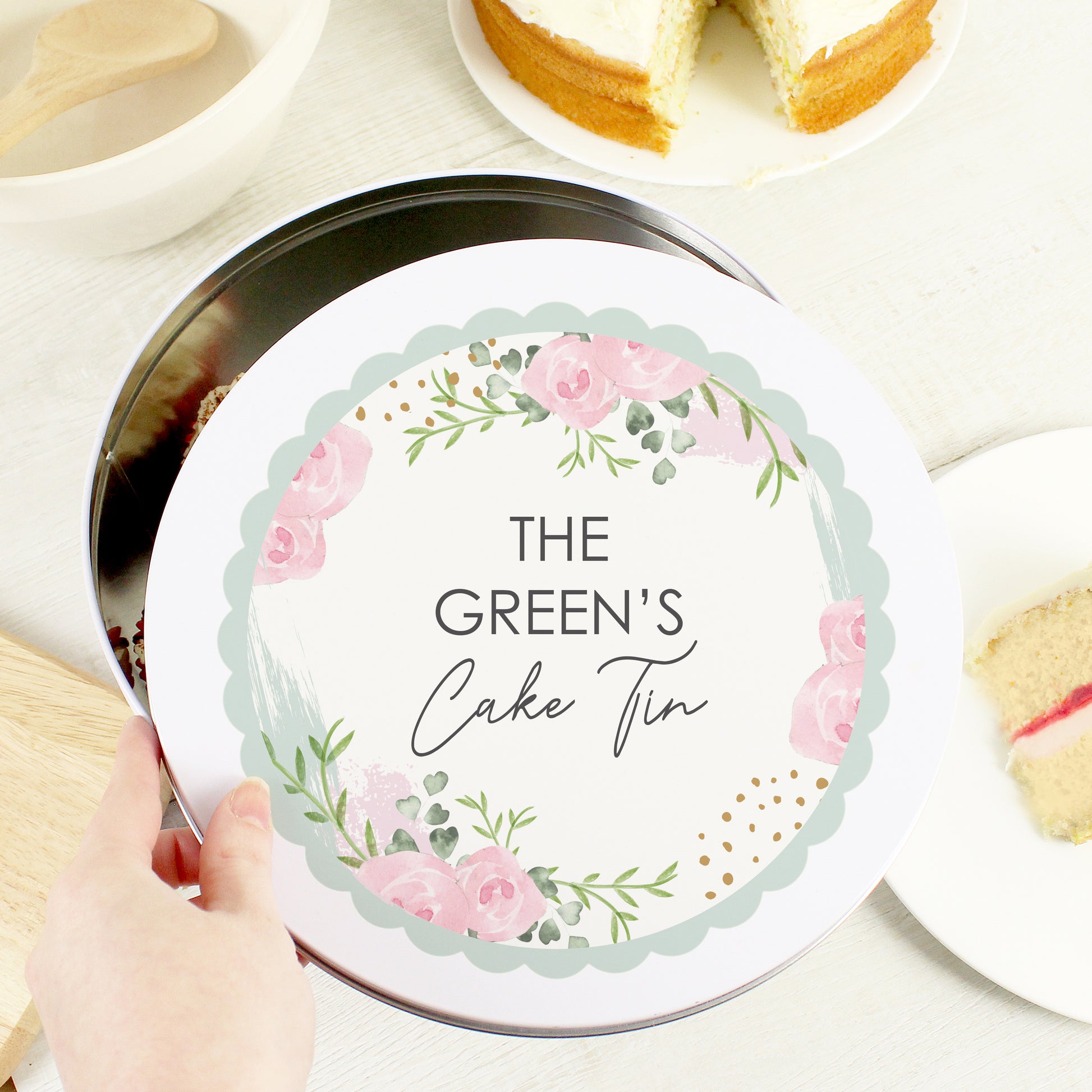 A hand is holding the tin cover of Personalised Floral Cake Tin that says "THE GREEN'S Cake Tin"