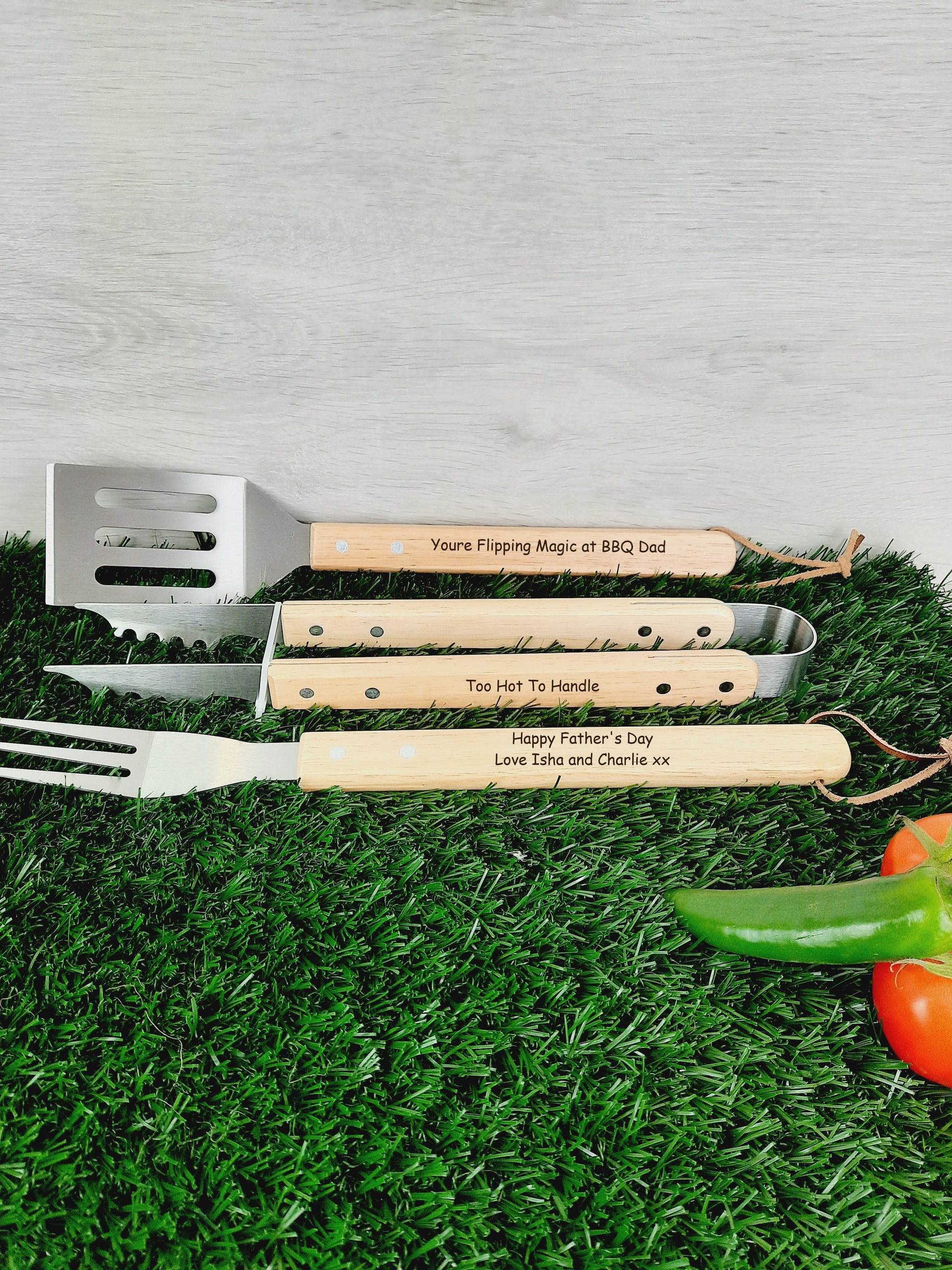 Gifts for BBQ Lovers lies on a faux grass with 1 green chili and 2 tomatoes. On the fork, the words "Happy Father's Day Love Isha and Charlie xx," on the tongs, "Too Hot To Handle," and on the spatula, "You're Flipping Magic at BBQ Dad " are engraved.