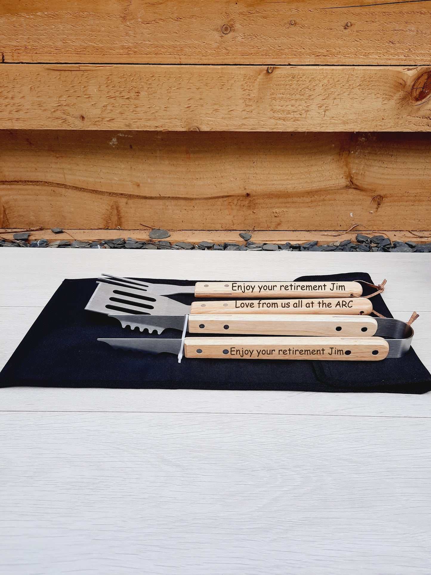 Personalised BBQ Tool Set lies in a black canvas bag. On the tongs, the words "Enjoy your retirement Jim," on the spatula, "Love from us all at the ARC," and on the fork, "Enjoy your retirement Jim" are engraved.
