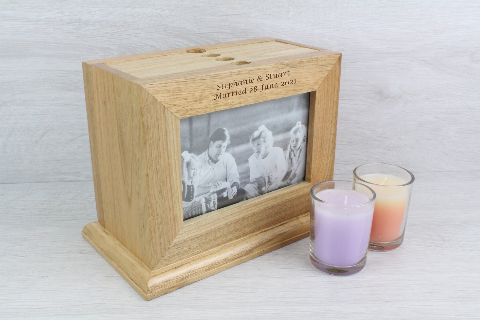 Sideview of a Personalised Oak Wooden Photo Frame and Album Holder with a text engraving that says "Stephanie & Stuart Married 28 June 2021" Inside the wooden photo frame, there's a black and white family photo with two young girls and their parents. Also, beside the wooden frame, there are two not lighted candles (colour purple and orange).